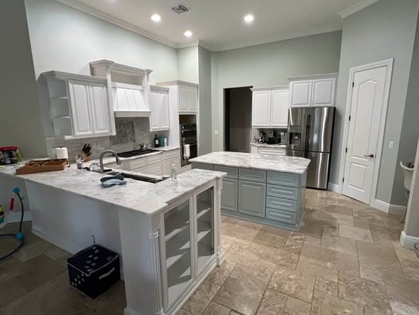 Wider view of the same two-tone kitchen — white perimeter cabinets with a soft green island and peninsula, marble counters and travertine floor