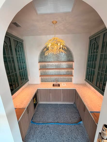 Butler's pantry mid-installation: arched glazed wall cabinets in deep green, fluted base units and fluted glass shelving, floor still protected